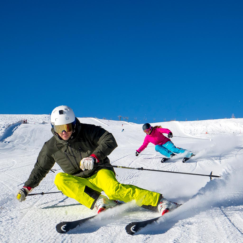 Nahaufnahme eines Mannes mit Skiern, der den Berg hinunterfährt, gefolgt von einer Frau im Hintergrund.