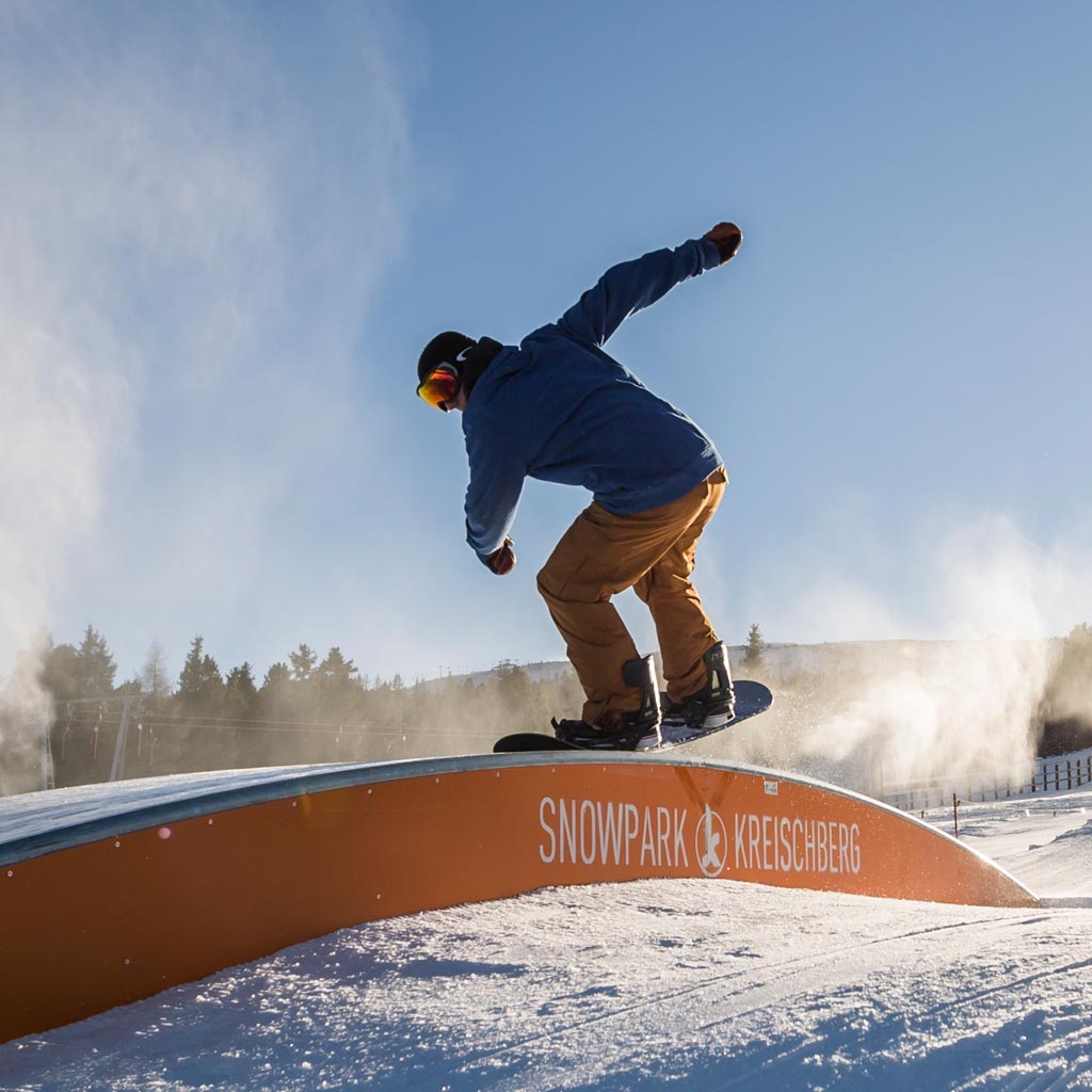 Skiboarder beim Sprung über ein Hindernis im Snowpark Kreischberg, mit Schriftzug 'Snowpark Kreischberg'