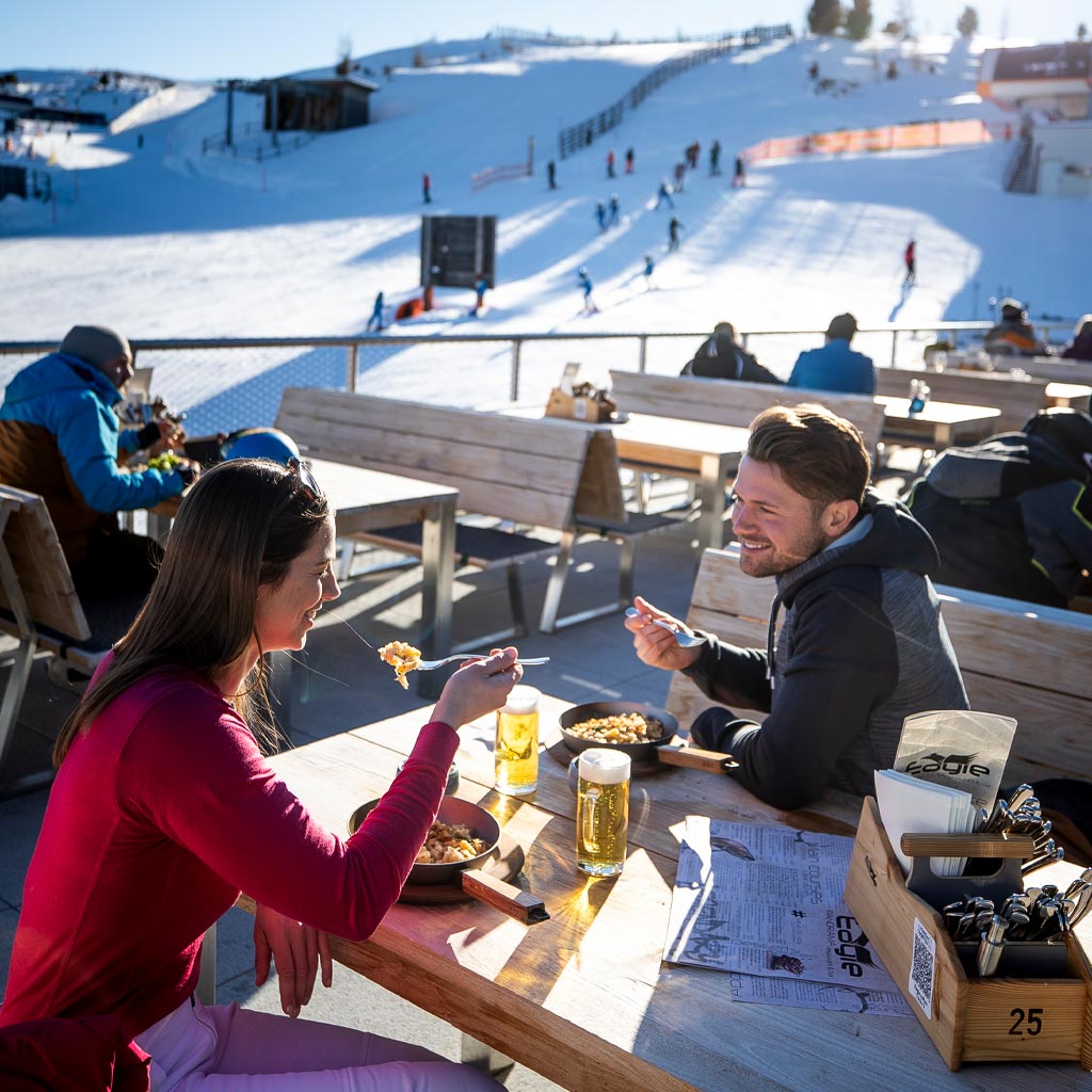 Glückliches Paar beim Essen im winterlichen Freien, mit Blick auf die Skipiste im Hintergrund.